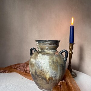 Op de afbeelding: Een keramische vaas met twee oren, voorzien van een gespikkeld glazuur in tinten bruin, beige en crème. De vaas heeft een brede, ronde vorm en een uitlopende rand. Daarnaast staat een koperen kandelaar met een brandende blauwe kaars.