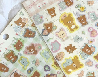 May include: Two sheets of sticker paper featuring a cute cartoon bear character. The stickers are on a checkered background with flowers and glitter accents. The text on the sticker sheets reads "Chairouikoguma".
