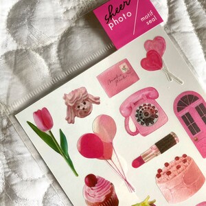 May include: Sheet of pink stickers with various designs, including a tulip, cupcake, lipstick, and telephone. The stickers are in a clear plastic sleeve with a pink label that reads "sheer photo motif seal."