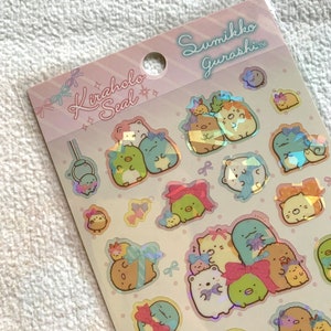 May include: A sheet of iridescent stickers featuring characters from the Sumikko Gurashi series. The stickers are on a pink and white background with the text "Kiraholo Seal" and "Sumikko Gurashi" in pink and white. The characters are all wearing bows and are in various poses.