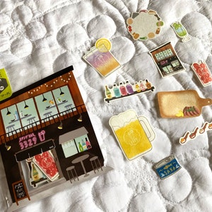 Peut inclure: Un sac en plastique transparent rempli d'autocollants colorés représentant des illustrations d'un bar, de boissons et de nourriture. Les autocollants sont de style japonais et comportent du texte en japonais. Le sac est posé sur une surface en tissu matelassé blanc.