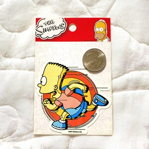 May include: A yellow and blue sticker featuring a cartoon character running in a circle. The character is wearing a red and blue shirt and blue pants. The sticker is labeled "The Simpsons" and "Matt Groening".