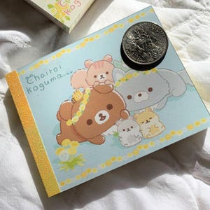 May include: A light blue notepad featuring cartoon bears and the text "Chairoi Koguma." The notepad has a yellow textured spine and a quarter coin on top. The illustrations include a brown bear, a gray bear, and smaller animal characters.