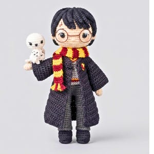 May include: A handcrafted crochet doll featuring a character with dark hair, round glasses, and a lightning bolt scar on the forehead. The doll wears a dark robe, a red and yellow striped scarf, and holds a small white owl on its right arm.