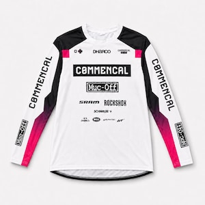 Peut inclure: Un maillot à manches longues blanc, noir et rose avec divers logos de marques dont Commencal, Muc-Off, Sram, Rockshox, Schwalbe, DHarco, Bell, E*thirteen et HT. Le haut présente un col rond et un motif dégradé rose sur les manches.