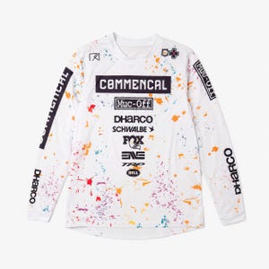 May include: A white long-sleeve jersey featuring a multi-coloured paint splatter pattern. The front displays the text COMMENCAL, Muc-Off, DHARCO, SCHWALBE, FOX, ENVE, TRP, and BELL. The sleeves feature the text COMMENCAL and DHARCO in black.