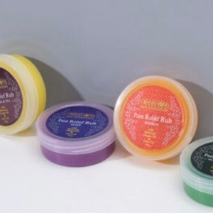 Puede incluir: Cuatro envases redondos de bálsamo para el alivio del dolor en amarillo, morado, naranja y verde. Cada envase tiene una etiqueta con el nombre del producto y los ingredientes. Los envases están sobre una superficie blanca.