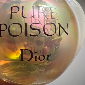 May include: A clear, iridescent perfume bottle with the words "PURE POISON" and "Dior" printed in black. The bottle has a rounded shape and a pearlescent sheen, reflecting light with a rainbow effect. The liquid inside is a golden color.