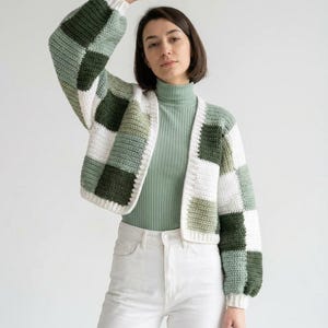 May include: A cropped crochet cardigan featuring a checkered pattern in shades of dark green, sage green, and white. The garment has long sleeves with ribbed cuffs, a button-front closure, and a V-neckline, worn over a sage green turtleneck.