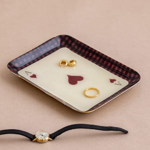 Può includere: Un vassoio rettangolare con il disegno di una carta da gioco asso di cuori e un bordo a scacchi rosso scuro e nero. Il vassoio contiene due piccoli orecchini a cerchio dorati e un sottile anello dorato. Sotto il vassoio c'è un orologio con cinturino nero.