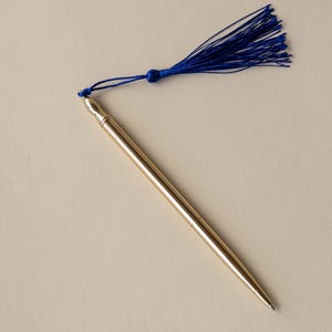 May include: A slim, metallic gold ballpoint pen featuring a smooth, cylindrical body and a pointed tip. Attached to the top of the pen is a vibrant royal blue thread tassel, secured by a small matching blue knot, creating a distinct contrast.