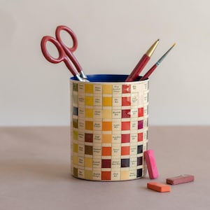 Könnte beinhalten: Ein zylindrischer Stiftehalter mit einem Raster aus verschiedenen Farbfeldern und aufgedruckten Textbeschriftungen. Im Halter befinden sich eine rote Schere, ein goldfarbener Stift und ein dünner Pinsel. Zwei kleine rechteckige rosa und orange Blöcke.