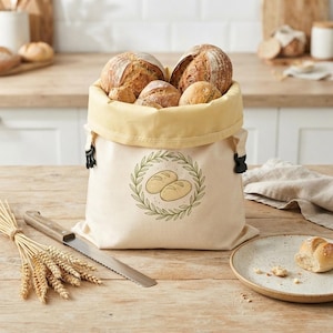 May include: A cream-colored canvas bread bag with a yellow interior lining and black side buckles. The front features a green laurel wreath graphic surrounding two illustrated loaves of bread. The bag is filled with several round, crusty loaves.