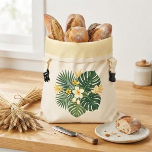 May include: A cream-colored canvas bread bag with a roll-top closure and black plastic side buckles. The front features a printed graphic of green monstera leaves and yellow plumeria flowers. The bag is filled with several crusty bread loaves.