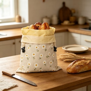May include: A beige fabric bread bag featuring a repeating pattern of white daisy flowers with yellow centers. The bag has a light yellow interior lining, a drawstring closure with black plastic toggles, and is filled with several crusty bread rolls.