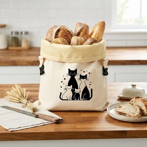 May include: A cream-colored canvas bread bag featuring a black graphic print of four cats of varying sizes. The bag has a wide, folded-over top edge and black side clips, and it is filled with several loaves of crusty artisan bread.