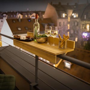 May include: A rectangular light wood balcony table shelf mounted on a railing. It holds a glass bottle, two stemmed wine glasses filled with white wine, a silver metal bowl with green salad, and a glass container filled with breadsticks.