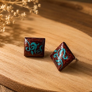 Peut inclure: Une paire de boucles d'oreilles à clous carrées présentant une finition texturée brun foncé semblable à du bois avec une surface brillante. Chaque boucle est ornée d'un motif calligraphique turquoise vif et tourbillonnant.