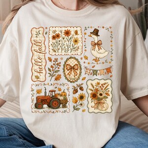 May include: A cream-colored t-shirt featuring a graphic print with the text "hello fall". The design includes a red tractor, a goose wearing a hat, sunflowers, pumpkins, acorns, and autumn leaves arranged in a collage of rectangular frames.