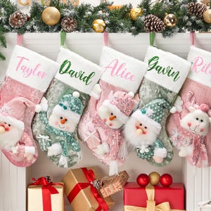 May include: Five personalised Christmas stockings hanging in a row. The stockings feature plush cuffs with the names Taylor, David, Alice, Gavin, and Riley embroidered in pink or green. Each stocking displays a festive character like a snowman or reindeer.