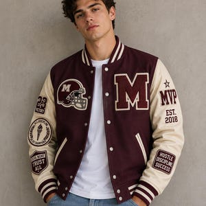 Op de afbeelding: Een bordeauxrood en crèmekleurig varsity-jack met een grote letter M op de borst, een afbeelding van een footballhelm en diverse patches waaronder MVP EST 2018, 23 VICTORY, ALL DISCIPLINE ALL CHAMPIONS, FEAR NONE TRUST ALL en HUSTLE DISCIPLINE SUCCESS.
