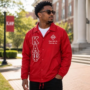 May include: A red windbreaker jacket featuring a white collar, snap button closure, and long sleeves. The front displays the Greek letters KΑΨ in white, with a diamond-shaped 1911 logo and the text Kappa Alpha Psi Fraternity, Inc. embroidered in white.