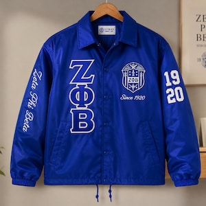 May include: A royal blue coach jacket featuring white embroidered Greek letters ZΦB on the front, a crest with the text Since 1920, and the number 1920 on the sleeve. The right sleeve displays the text Zeta Phi Beta in white script lettering.