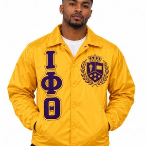 May include: A bright yellow snap-front coach jacket featuring a vertical purple and white Greek letter design on the right chest and a purple and white crest emblem on the left chest, including a crown, torch, book, and stars.