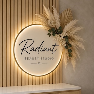 Peut inclure: Une enseigne murale circulaire rétroéclairée portant le texte Radiant BEAUTY STUDIO avec une petite icône en forme de cœur. L'enseigne est ornée d'un arrangement décoratif d'herbes de la pampa séchées, de feuilles de palmier et d'eucalyptus.