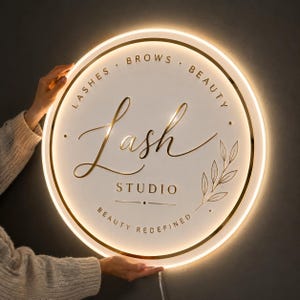 Może przedstawiać: Okrągły biały szyld ścienny ze złotym napisem i grafiką złotych liści. Tekst brzmi LASHES BROWS BEAUTY, Lash STUDIO oraz BEAUTY REDEFINED. Szyld posiada świecący ciepłym światłem pierścień LED wokół krawędzi oraz przewód zasilający u dołu.