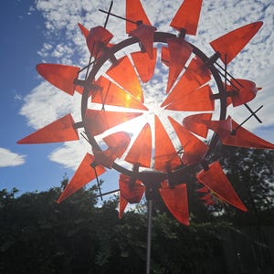 May include: A circular garden wind spinner featuring numerous translucent orange, triangular-shaped plastic blades attached to a dark metal frame. The structure is mounted on a thin metal pole, with the sunlight shining through the orange blades.
