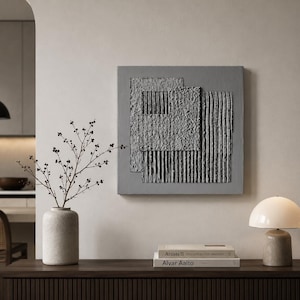 May include: A square gray wall art piece featuring a textured composition of overlapping geometric panels with varying surface patterns, including rough stucco-like sections and vertical ribbed lines, mounted on a flat gray background.