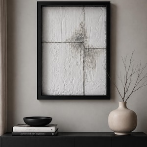 May include: A rectangular wall art piece with a black frame, featuring a white textured surface divided into four panels with dark gray weathering. Below, a black bowl sits on two stacked books, next to a beige vase with thin bare branches.