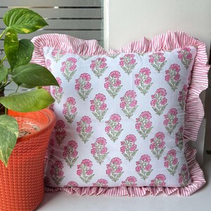 Könnte beinhalten: Ein quadratisches Zierkissen mit weißem Grund und einem sich wiederholenden rosa Blumenmuster mit grünen Stielen. Das Kissen ist von einem gerüschten Rand aus rosa-weiß gestreiftem Stoff umgeben, der eine dekorative Kante bildet.