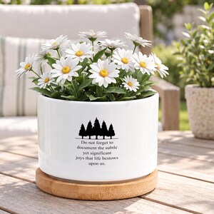 May include: A white cylindrical ceramic planter with a bamboo saucer base, containing a cluster of white daisies with yellow centres. The planter features a black graphic of pine trees and the text: Do not forget to document the subtle yet significant joys that life bestows upon us.