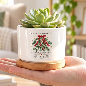 May include: A white ceramic planter with a bamboo base containing a green succulent. The planter features a Christmas mistletoe graphic with a red bow and the text: OUR FIRST CHRISTMAS AT 2567 MAPLE GROVE LANE, Amanda & Eric, 2026.