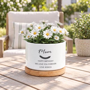 Pode incluir: Um vaso cilíndrico de cerâmica branca contendo um buquê de margaridas brancas com centros amarelos. O vaso apresenta texto em preto que diz "Mom", "HAPPY BIRTHDAY", "WE LOVE YOU FOREVER" e "LOVE JESSICA" acima de um gráfico de folhas.