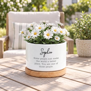 Pode incluir: Um vaso cilíndrico de cerâmica branca com uma base de bambu, contendo um grupo de margaridas brancas com centros amarelos. O vaso apresenta o nome Sophia e o texto: Some people just make the world a better place. You are one of those people.
