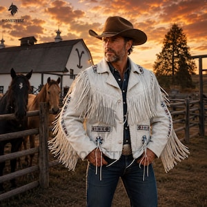 May include: A cream-coloured suede jacket featuring long fringe detailing on the chest and sleeves, accented with intricate geometric beadwork patterns in blue, red, and black, worn over a dark button-down shirt and blue denim jeans.