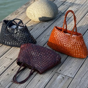 May include: Three woven tote bags in black, dark brown, and tan. Each bag features a structured, rectangular base, a wide opening, and two braided handles. The material has a consistent, interlocking grid pattern throughout the entire exterior.