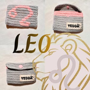 May include: A gray crochet pouch featuring a pink Leo zodiac symbol on the front. The pouch has a pink snap closure and a white fabric label with the text VEDDA CROCHET stitched on the front. The image includes the text LEO and a lion graphic.