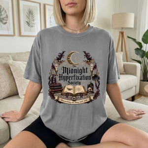 May include: A grey t-shirt featuring a graphic print with the text The Midnight Hyperfixation Society and Researching one thing until 3AM. The design includes a crescent moon, an open book, a skull, a potion bottle, and moths.