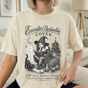 Puede incluir: Una camiseta beige con un gráfico negro de una bruja leyendo un libro, un gato, una lista de tareas y una casa. El texto dice: Executive Dysfunction COVEN y All Good Intentions Welcome MEETS EVENTUALLY, PROBABLY, MAYBE.