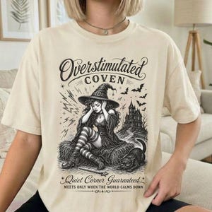 May include: A cream-colored t-shirt featuring a black graphic print of a witch in a pointed hat sitting on a haystack with a cat, a haunted house, and bats. The text reads Overstimulated Coven and Quiet Corner Guaranteed, Meets Only When The World Calms Down.