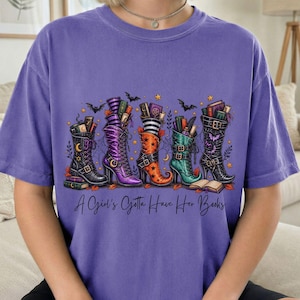 May include: A purple t-shirt featuring a graphic of four tall lace-up boots filled with books. The boots are black, purple, orange, and green, decorated with stars and bats. The text "A Girl's Gotta Have Her Books" is printed in black script below.