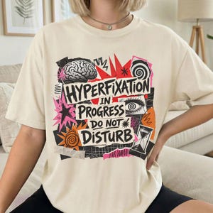 May include: A cream-colored short-sleeve t-shirt featuring a graphic print with the text "HYPERFIXATION IN PROGRESS DO NOT DISTURB". The design includes a brain, a spiral, an eye, an hourglass, and star shapes in black, red, pink, and orange.