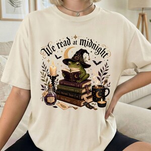 May include: A cream-coloured t-shirt featuring a graphic of a green frog wearing a witch hat and reading a book atop a stack of books. The design includes a candle, a potion bottle, a mug, and the text "We read at midnight" in black script.