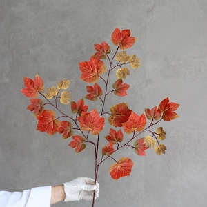 May include: A decorative artificial branch featuring multiple maple-style leaves in vibrant shades of red, orange, and gold. The branch has a thin, dark brown stem that splits into several smaller twigs, each adorned with textured, multi-coloured foliage.