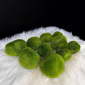May include: A collection of eleven round, textured moss balls in varying shades of vibrant green. Each sphere features a dense, fuzzy surface that mimics the appearance of natural moss, arranged in a cluster on a white faux fur surface.