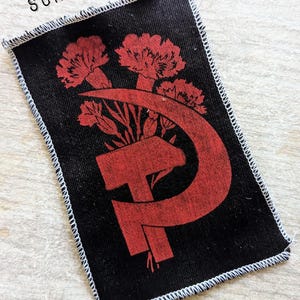 Peut inclure: Un écusson rectangulaire en tissu noir avec un bord surjeté blanc. L'écusson présente une impression graphique rouge représentant une faucille et un marteau entrelacés avec trois œillets. Le texte WHITE OVERLOCK SURJET BLANC est visible.